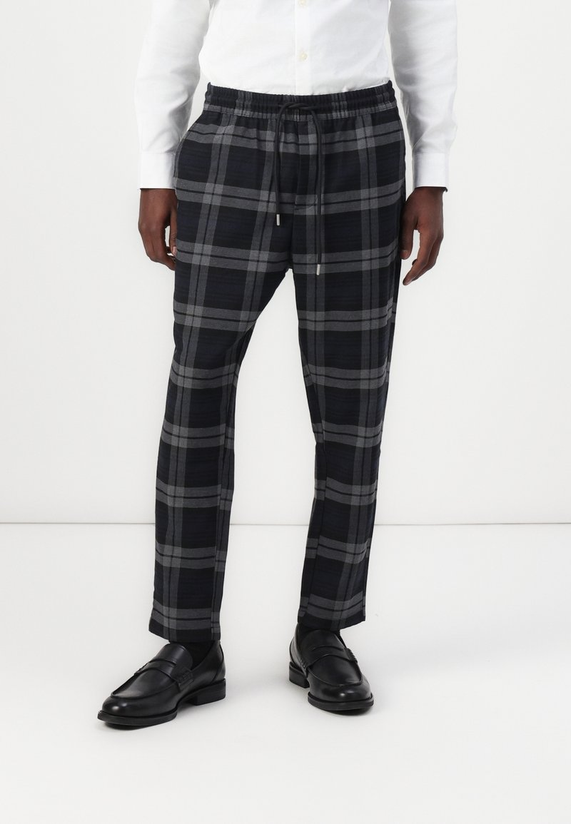 Pantalon à carreaux noir et gris avec une taille élastique et un cordon de serrage. Le tissu a une texture lisse. Associé à des chaussures en cuir noir.