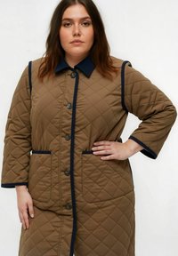 Manteau matelassé marron avec un col, bordé de marine, et une fermeture à boutons. Comprend deux poches avant et des manches trois-quarts.