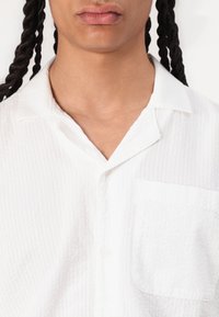 Chemise blanche à texture, à col à revers, avec une poche poitrine simple et un tissu léger, présentant de subtiles rayures horizontales.