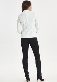 Blazer bianco aderente con design strutturato, maniche lunghe, cuciture sottili, abbinato a jeans skinny neri e scarpe con tacco alto.