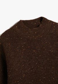 Bruine gebreide trui met een geribbelde textuur. Heeft een hoge, ronde halslijn en subtiele spikkels van een lichtere kleur door de stof.