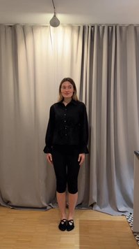 Γυναίκα στέκεται μέσα σε ξύλινο πάτωμα μπροστά από μπεζ κουρτίνες, φοράει μαύρο πουκάμισο με κουμπιά, κοντά παντελόνια και μαύρα παπούτσια τύπου slip-on.