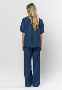 Denim dviejų dalių komplektas, kuriame yra laisvo kirpimo viršus su pūstomis rankovėmis ir plačios kelnes, abi tamsiai mėlynos su tekstūruotu finišu.