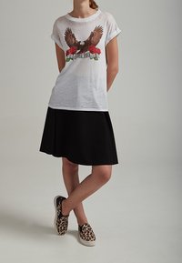 Hvid grafisk t-shirt med ørner og roser design, parret med en sort knælang nederdel og slip-on sko med leopardprint. Glattere stofstruktur.