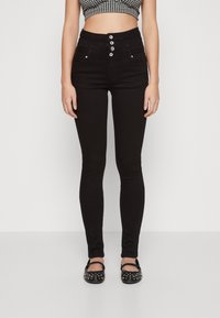 Jeans skinny negros de tiro alto con cierre de cinco botones, bolsillos frontales y textura suave; llevados con flats negros con tachuelas.