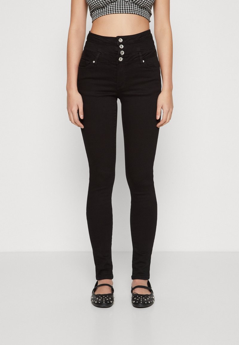 Jeans skinny negros de tiro alto con cierre de cinco botones, bolsillos frontales y textura suave; llevados con flats negros con tachuelas.