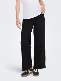 Persona valkā melnas plaša piegriezuma džinsas, melnas un baltas sporta kurpes un baltu T-kreklu, stāvot pret vienkāršu gaišu fonu.