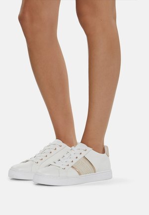 Sneakers basses blanches avec mesh beige et accents métalliques dorés portées sur des jambes nues devant un fond uni.