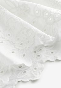 Balts izšūts audums ar ziedu rakstiem un viļņotām malām, ar izgriezuma detaļām un mīkstu, teksturētu virsmu.