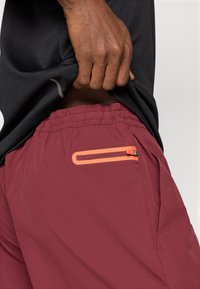 Bordové športové šortky s elastickým pásom, obsahujúce bočnú vrecko s oranžovým zipsom. Hladká, ľahká textúra látky.