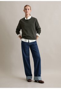 Mörkgrön tröja över en vit skjorta med krage, matchat med lösa blå jeans med uppvikta benslut. Bruna klackboots kompletterar outfiten.