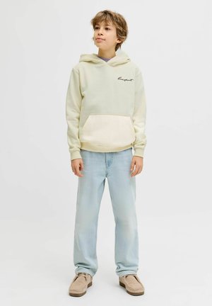 Poika, jolla on päällään kerma vaalea huppari, jossa etutasku, vaaleansiniset farkut ja vaaleanruskeat rennot kengät, seisoo yksinkertaisen valkoisen taustan edessä.