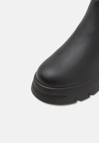 Botte en caoutchouc noire avec une semelle épaisse à nervures et un bout rond lisse, présentée sur un fond clair uni.