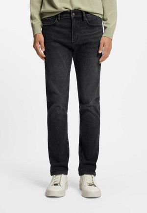 Uomo che indossa jeans neri skinny e sneakers bianche in piedi su uno sfondo chiaro e uniforme.