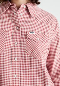 Chemise à carreaux rouges et blancs avec deux poches avant, un col et une petite étiquette "Wrangler". Le tissu semble léger et texturé.
