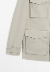 Rukáv světle béžové bundy s dvěma velkými klopovými kapsami a detailem nastavitelného pásku na manžetě na bílém pozadí.