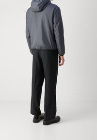 Graue leichte Jacke mit Kapuze, elastischem Taillenbund und langen Ärmeln, kombiniert mit schwarzen weit geschnittenen Hosen und schwarzen Schuhen.
