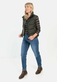 Olivgrüne Daunenweste mit hohem Kragen, über einem navy- und braun gestreiften Hemd getragen, kombiniert mit blauen Jeans und braunen Schnürstiefeln.