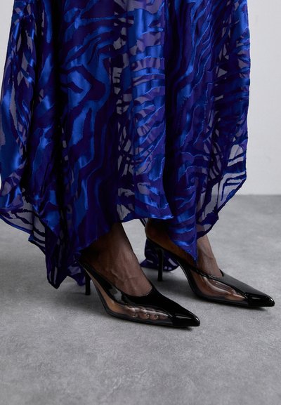 Black patent leather high-heeled shoes featuring a pointed toe and sheer side panels, paired with a flowing blue patterned dress.