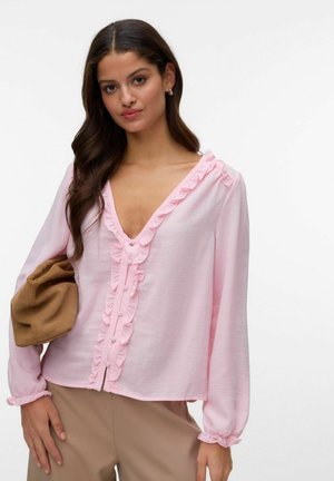 Femme aux longs cheveux bruns portant un chemisier rose pâle à volants et un pantalon beige, tenant une pochette marron pliée sous un bras.