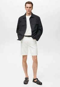 Schwarze Button-Up-Jacke mit zwei Fronttaschen, weißem T-Shirt, weißen Shorts und schwarzen Sandalen. Einfaches Design, gerade Passform und leichter Stoff.