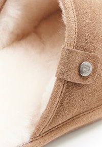 Pantofole in suede marrone con una soffice fodera interna beige chiaro. Presentano un dettaglio a bottone argentato e cuciture decorative. Forma arrotondata.