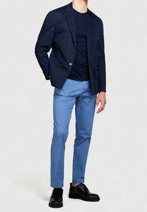 Hombre con blazer azul marino, camiseta negra, pantalones azul claro y zapatos de vestir negros, de pie frente a un fondo liso.