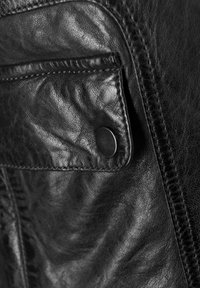 Nærbillede af en lomme på en sort læderjakke med synlig struktur, syninger og en rund metalknap.