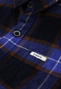 Kostkovaná košile v tmavě modré, hnědé a bílé barvě. Obsahuje knoflíky, límec a štítek "Garcia" na kapsičce. Texturovaná tkanina.