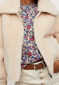 Persona vistiendo una chaqueta esponjosa de color crema sobre una camisa con estampado floral de flores rojas, azules y moradas, pantalones beige y un cinturón marrón.