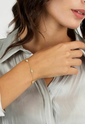 Bracciale in oro con tre accenti sferici, caratterizzato da una superficie texturizzata che scintilla, indossato su un braccio di tonalità chiara contro una camicia di satin.