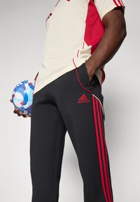 Schwarze Adidas-Trainingshosen mit roten Akzenten und drei roten Streifen. Cremfarbener Hemd-Detail sichtbar, der einen blau-weißen Fußball hält.