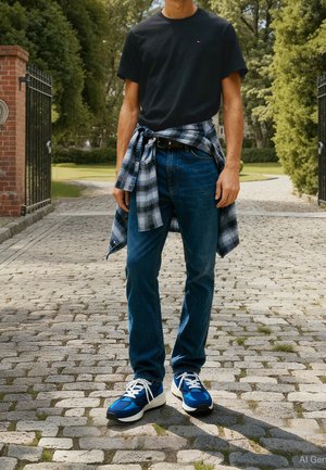 Man wearing blue jeans, blue and white sneakers, black t-shirt, and plaid shirt tied around waist standing on a cobblestone path outdoors.
