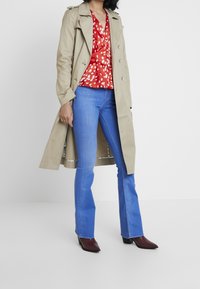 Trench beige con cierre de botones, blusa con estampado rojo y blanco, jeans acampanados azules y botas burdeos de punta.