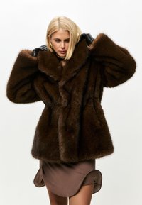 Brun faux fur frakk med en bred krave og voluminøse ærmer. Har en glat tekstur og er sat sammen med en tætsiddende brun nederdel.