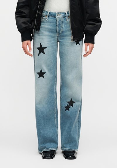 Jean délavé bleu clair avec des patchs étoiles noires sur l'avant des cuisses et des genoux, porté avec des chaussures noires et une veste noire.