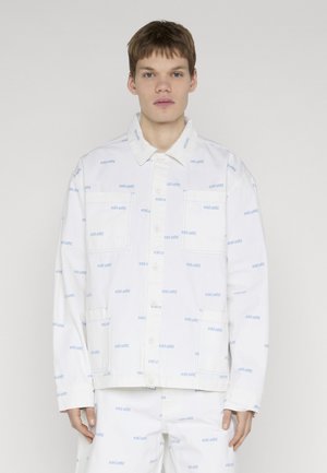 Mladi muškarac u bijeloj košulji s gumbima i hlačama s printom svijetloplavog teksta "eckō unltd." na jednostavnoj bijeloj pozadini.