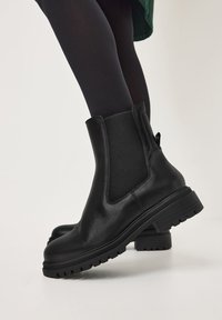 Botas de piel negras hasta el tobillo con paneles elásticos en los laterales, combinadas con medias opacas negras sobre un fondo blanco.