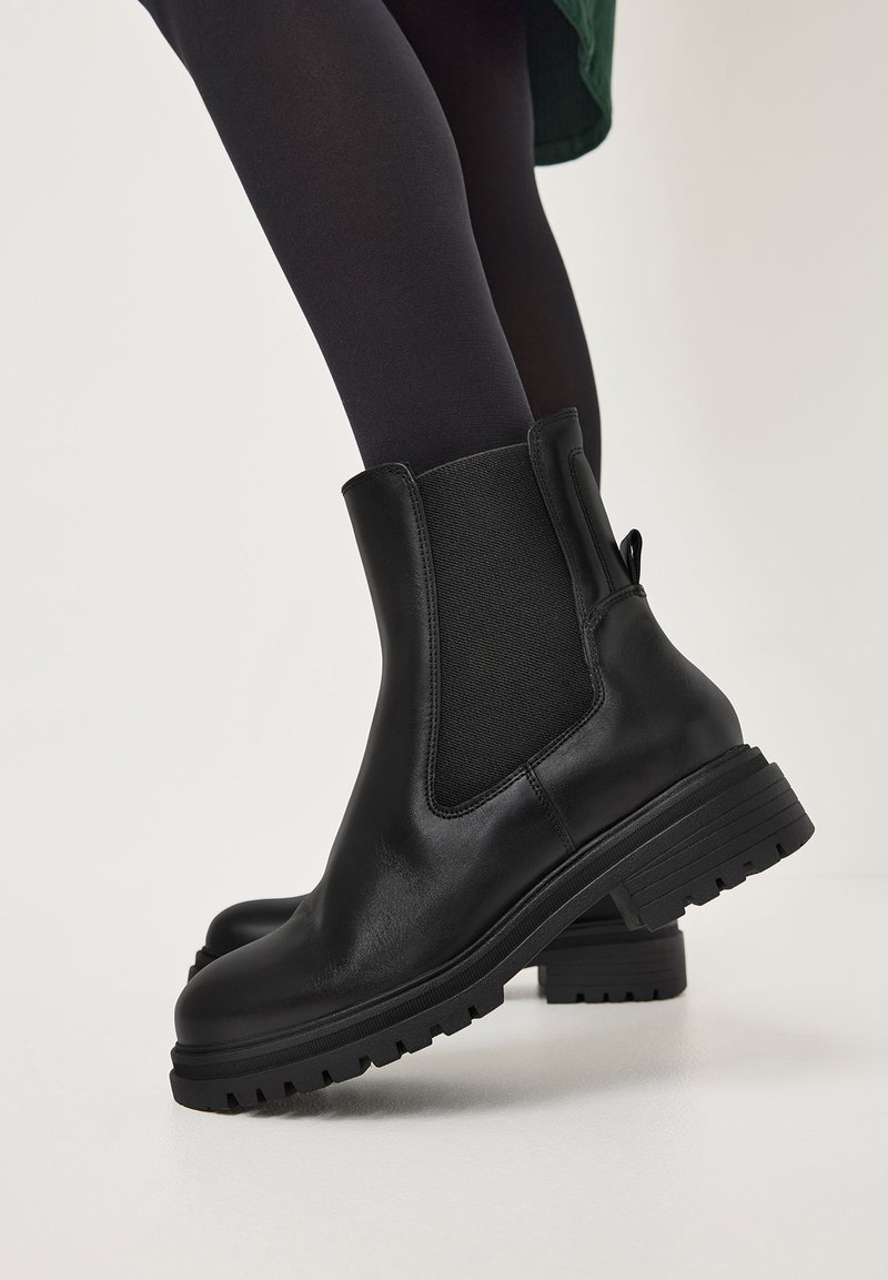 Botas de piel negras hasta el tobillo con paneles elásticos en los laterales, combinadas con medias opacas negras sobre un fondo blanco.