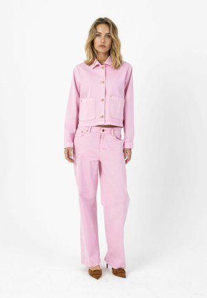 Lichtroze spijkerjack met twee steekzakken aan de voorkant, knoopsluiting en bijpassende lichtroze wijde jeans. Bruine hakken maken de outfit compleet.