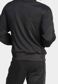 Svart sportjacka i slät tyg, med hög krage, dragkedja i ryggen och texturerade designdetaljer på axlarna.