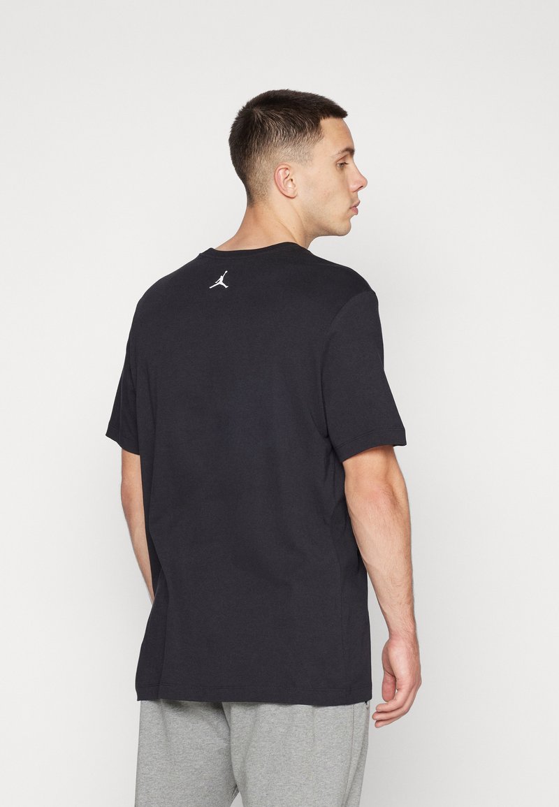 Hombre de perfil viste una camiseta negra de Jordan con un logo blanco en la parte superior de la espalda y pantalones de chándal gris claro contra un fondo blanco.