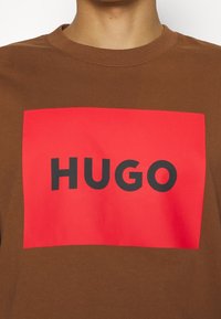 T-shirt en coton marron avec un grand graphisme rectangulaire rouge portant le mot "HUGO" en lettres noires en gras. Col rond et manches courtes.