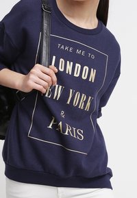 Felpa blu navy con tipografia dorata che riporta le parole "Londra", "New York" e "Parigi". Presenta un collo rotondo, maniche corte e orlo rifinito.