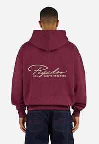 Burgundy hoodie with a front pocket and drawstring hood, features a cream logo text, paired with dark denim jeans with contrasting stitching.