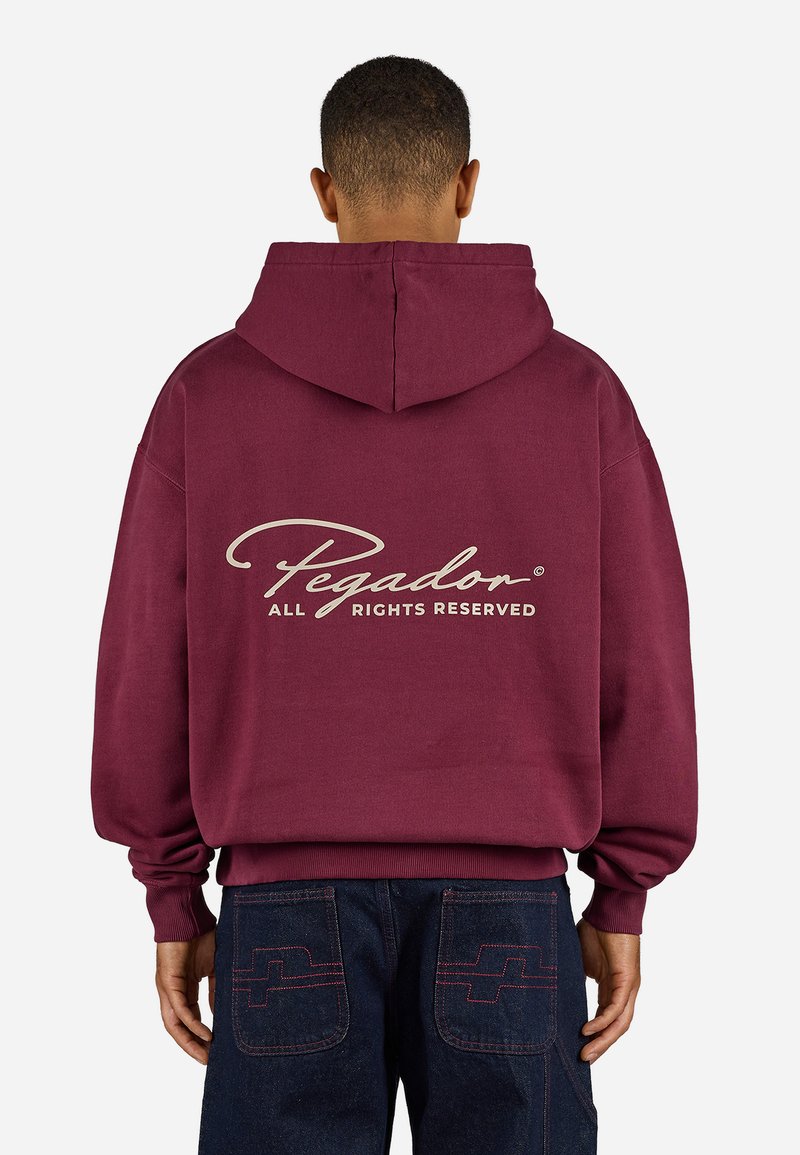Burgundy hoodie with a front pocket and drawstring hood, features a cream logo text, paired with dark denim jeans with contrasting stitching.