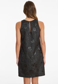 Frau in einem ärmellosen schwarzen Kleid mit glänzenden Blumen- und Wirbelmustern, von hinten vor einem weißen Hintergrund gesehen.