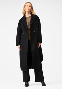 Sort uld lang frakke med slagkrave, dobbeltknappede knapper og oversized silhuet. Stylet med sorte bukser og hæle.