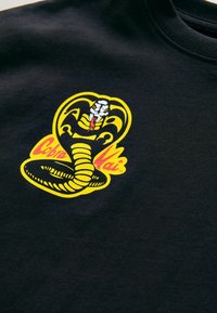 Sort bomulds t-shirt med et farverigt kobra grafisk design i gul, hvid og rød, med indviklede detaljer og et fremtrædende slange-design.