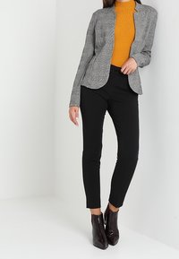 Una donna posa indossando un blazer grigio aderente su un dolcevita arancione e pantaloni neri, con stivaletti lucidi su uno sfondo neutro.